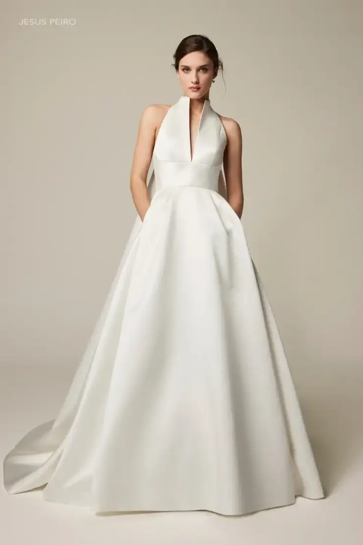 A woman in an elegant, white satin halter-neck gown with a deep V-neckline poses confidently against a neutral background, exuding sophistication.