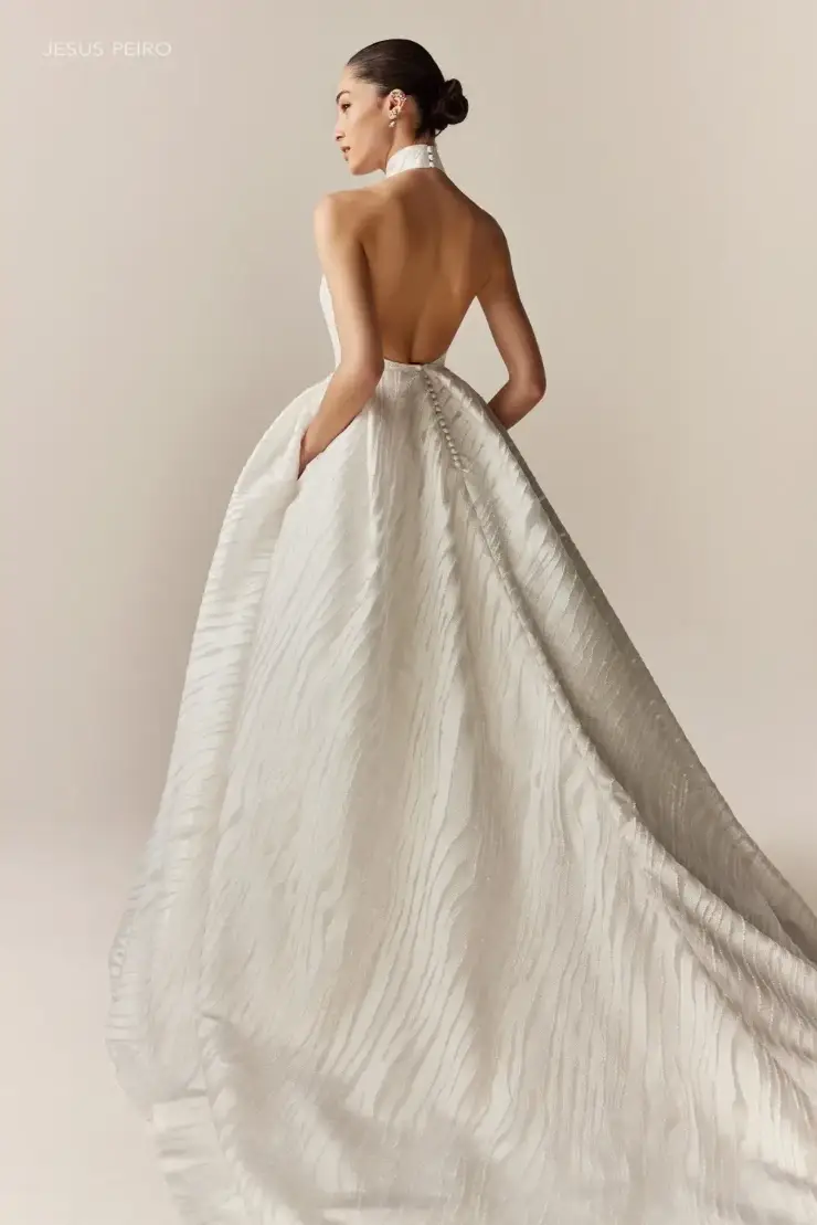 A woman in a flowing, textured white gown with a halter neck and open back, standing elegantly against a beige background. Her hair is styled in a bun.