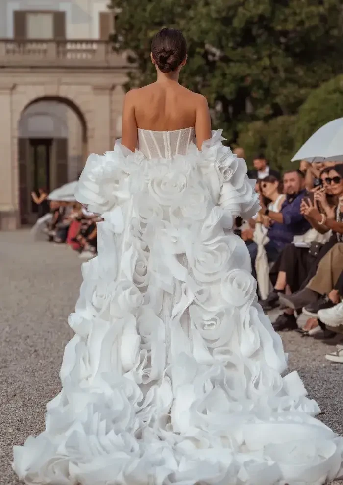A model walks outdoors wearing an elaborate white gown with large, floral fabric details. The audience on either side watches intently, capturing the moment.