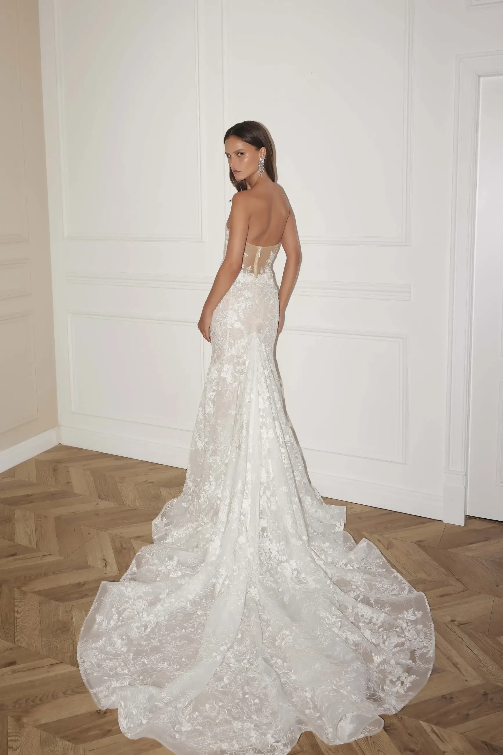 A woman in a strapless, lace wedding gown with a long train stands on a wooden floor, facing away. The atmosphere is elegant and serene.