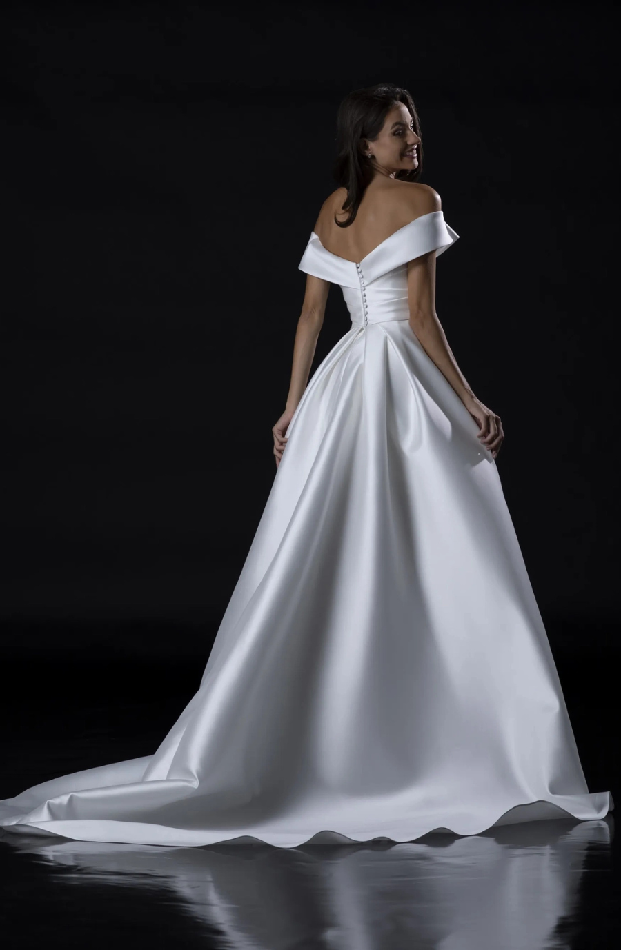 A woman in an elegant, off-the-shoulder white gown with a flowing train stands against a dark background, exuding a timeless and graceful elegance.