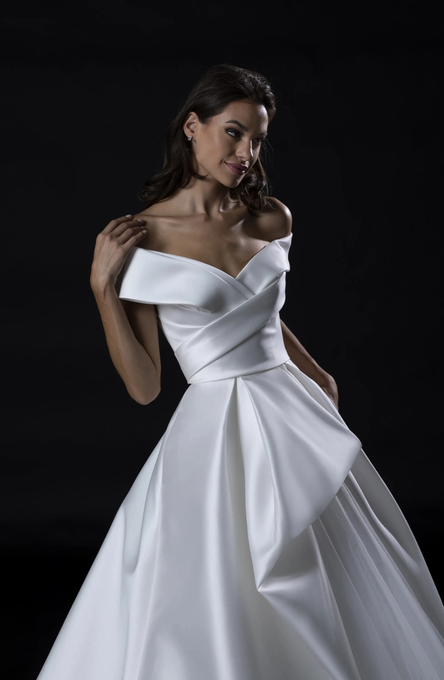 A woman in an elegant off-the-shoulder white wedding gown stands against a dark background. The dress features a fitted bodice and flowing skirt, exuding sophistication.