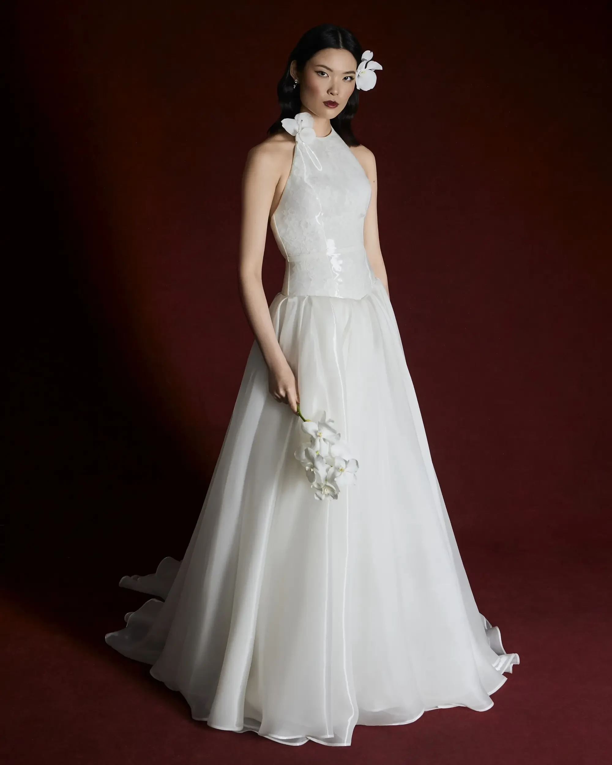 A woman in a flowing white wedding dress stands against a deep burgundy background. She holds white flowers, with serene expression, exuding elegance.