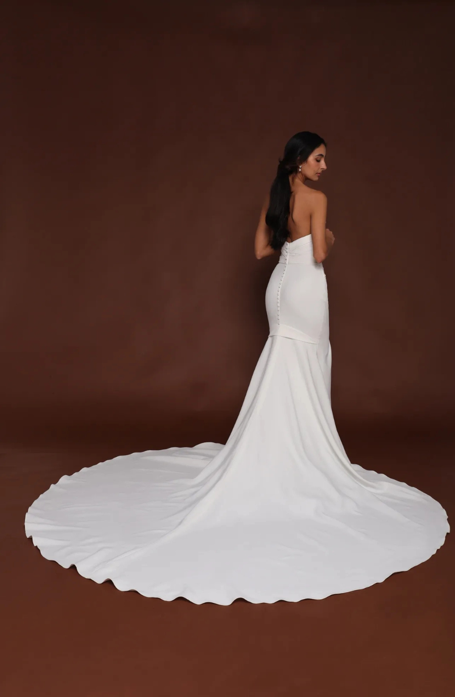 Model is wearing a strapless mermaid gown in 4-ply crepe with a banded bodice, half-circle godet skirt, cathedral train, and covered buttons.