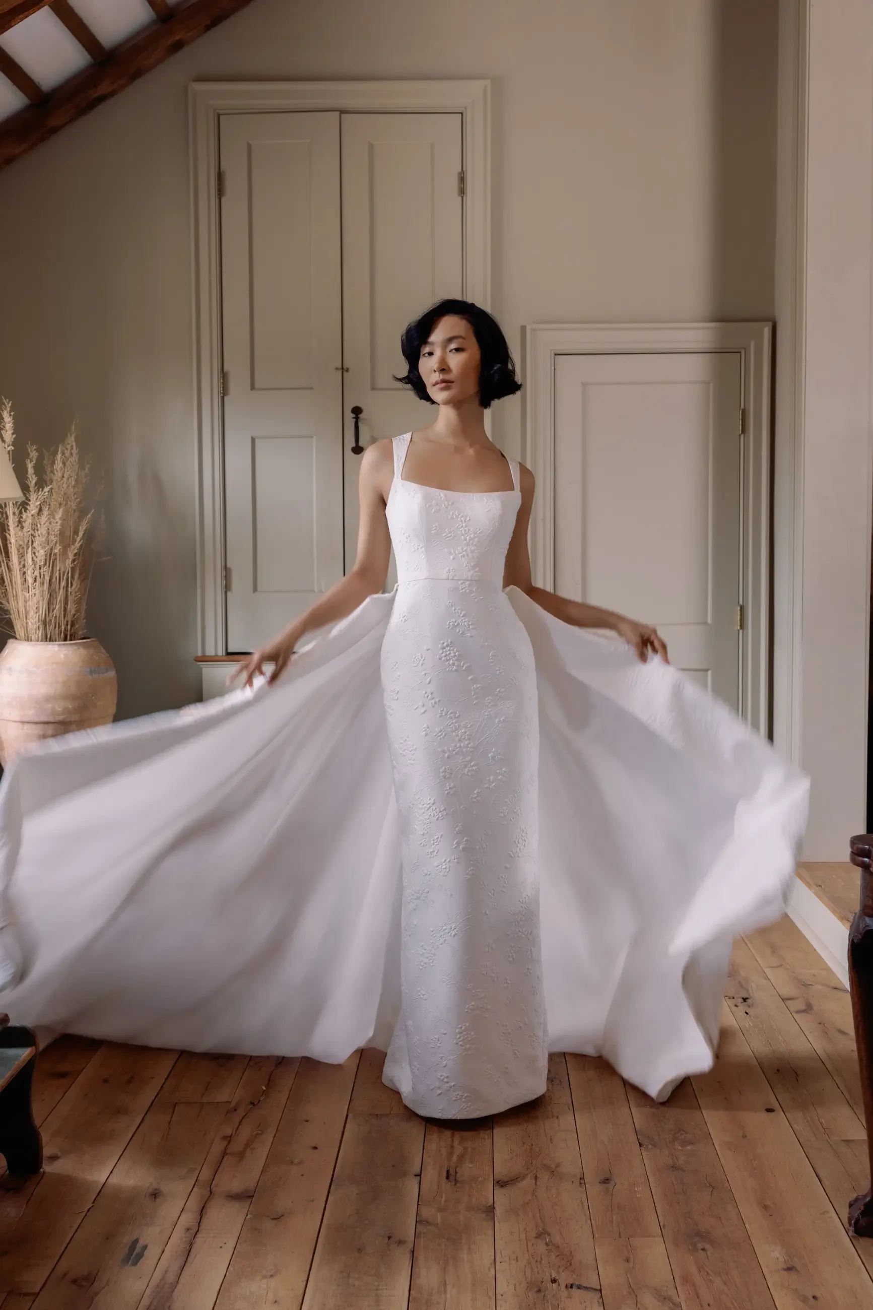 A person in a white dress stands elegantly in a room with wooden flooring, showcasing the flowing fabric of the dress.