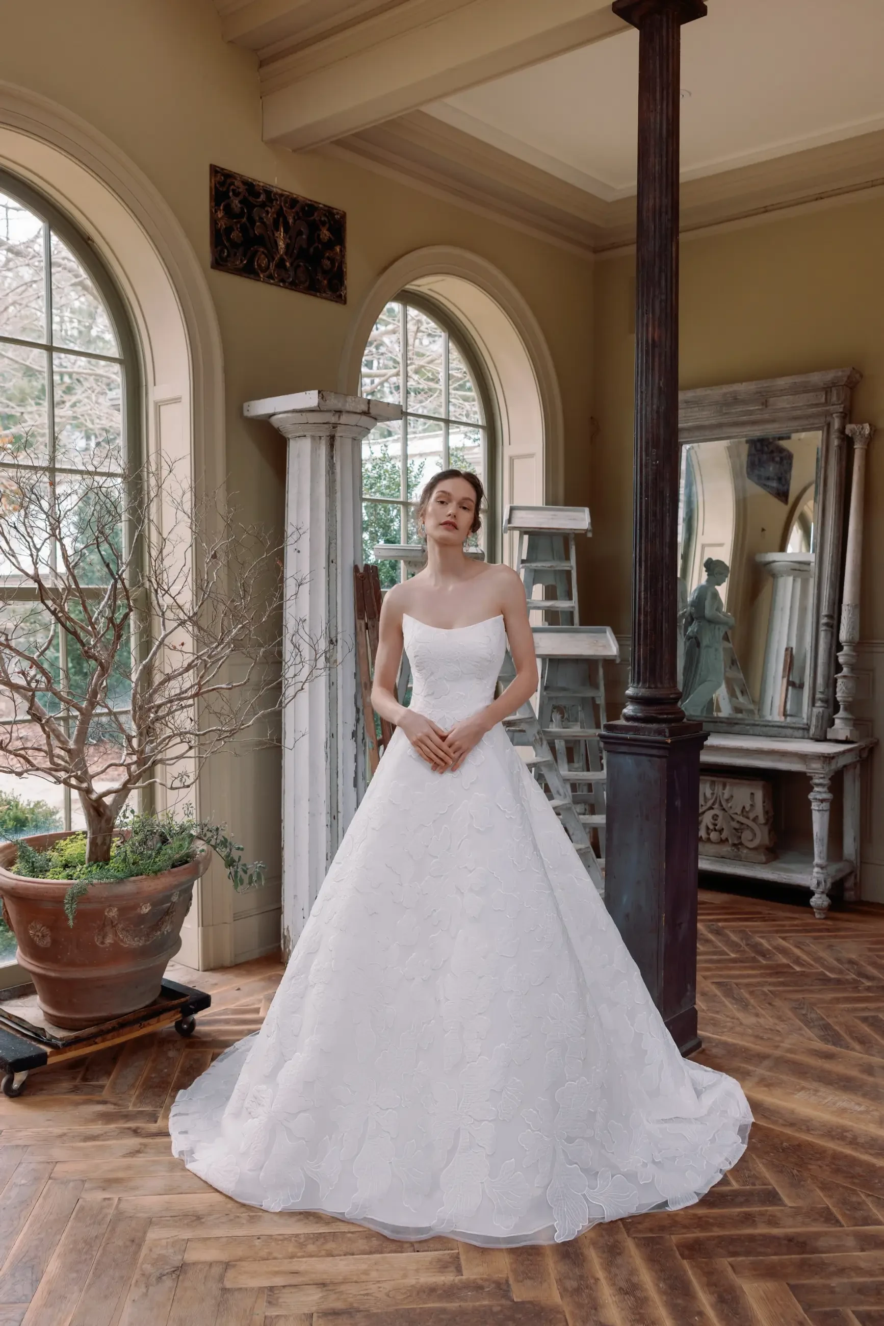 A woman wearing a white wedding dress stands in an elegantly decorated room featuring large windows, wooden flooring, and classic furnishings.