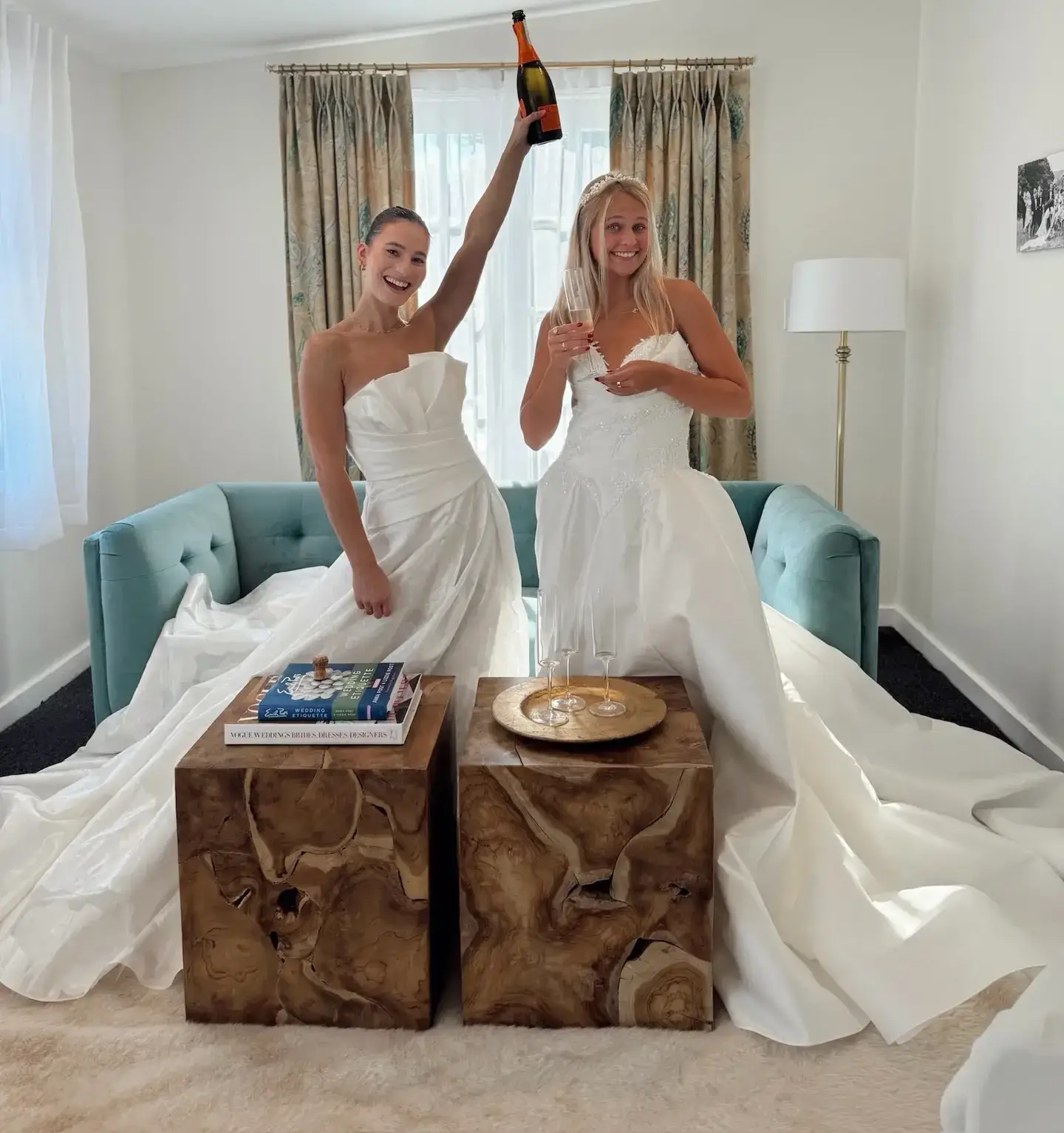Two women in white dresses are standing on a couch, one is holding up a champagne bottle while the other is holding a glass. They are celebrating, and there are decorative tables beside them.