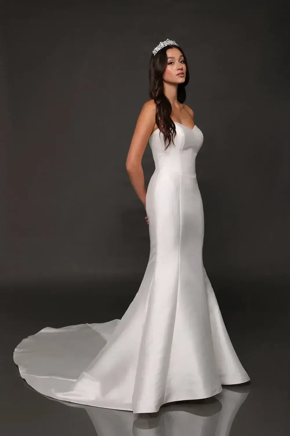 A model poses in a white wedding gown with a fitted silhouette and a long train, wearing a tiara.