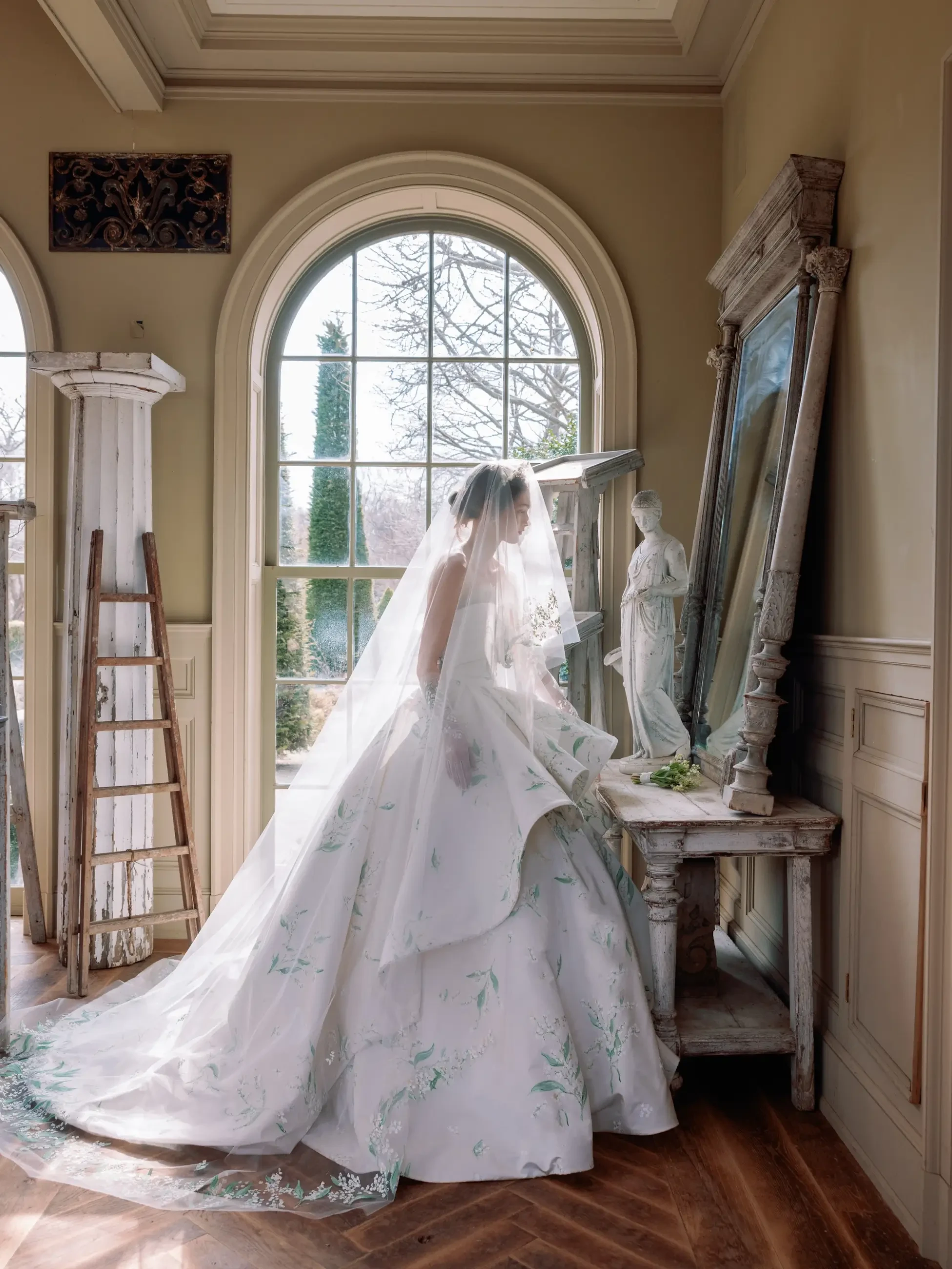 A bride in a floral wedding dress stands by a mirror, adjusting her veil in a softly lit room with large windows.