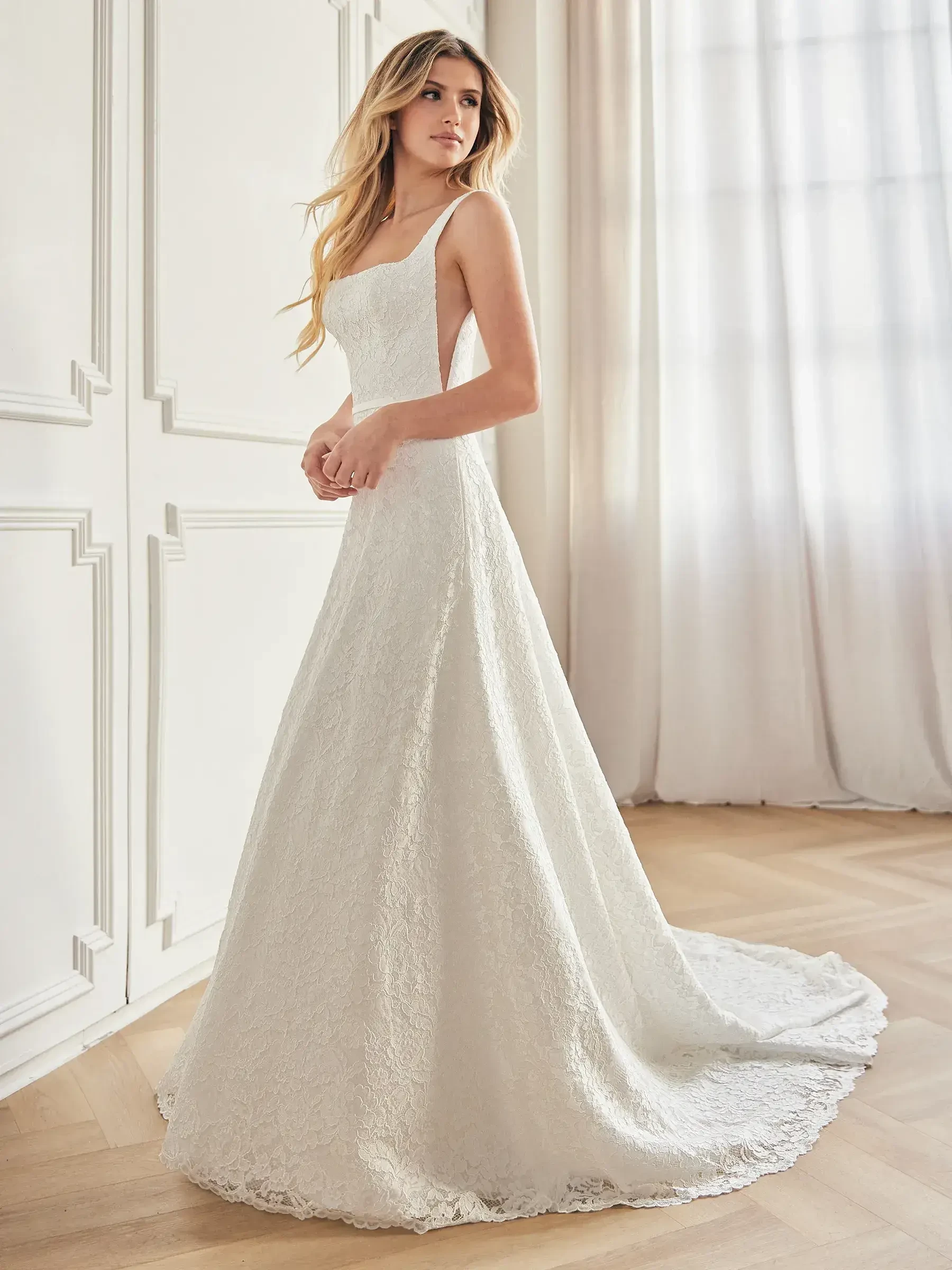 Bride in lace gown stands by window, exuding elegance. The dress features a fitted bodice and flowing train. Soft, natural light fills a classic room.