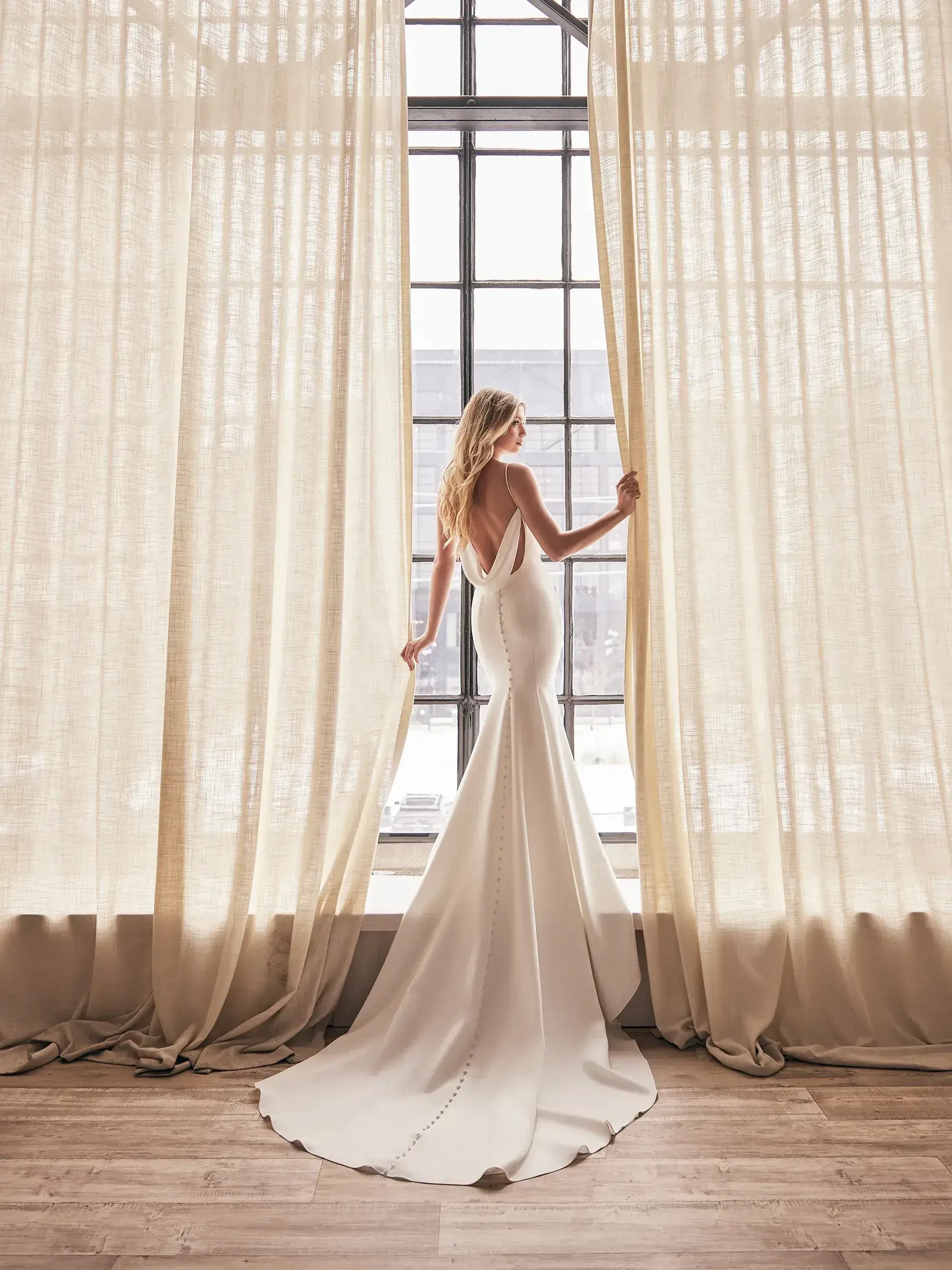 A woman in an elegant, backless white gown stands between sheer beige curtains, gazing out a large window. The scene evokes a sense of serenity and anticipation.