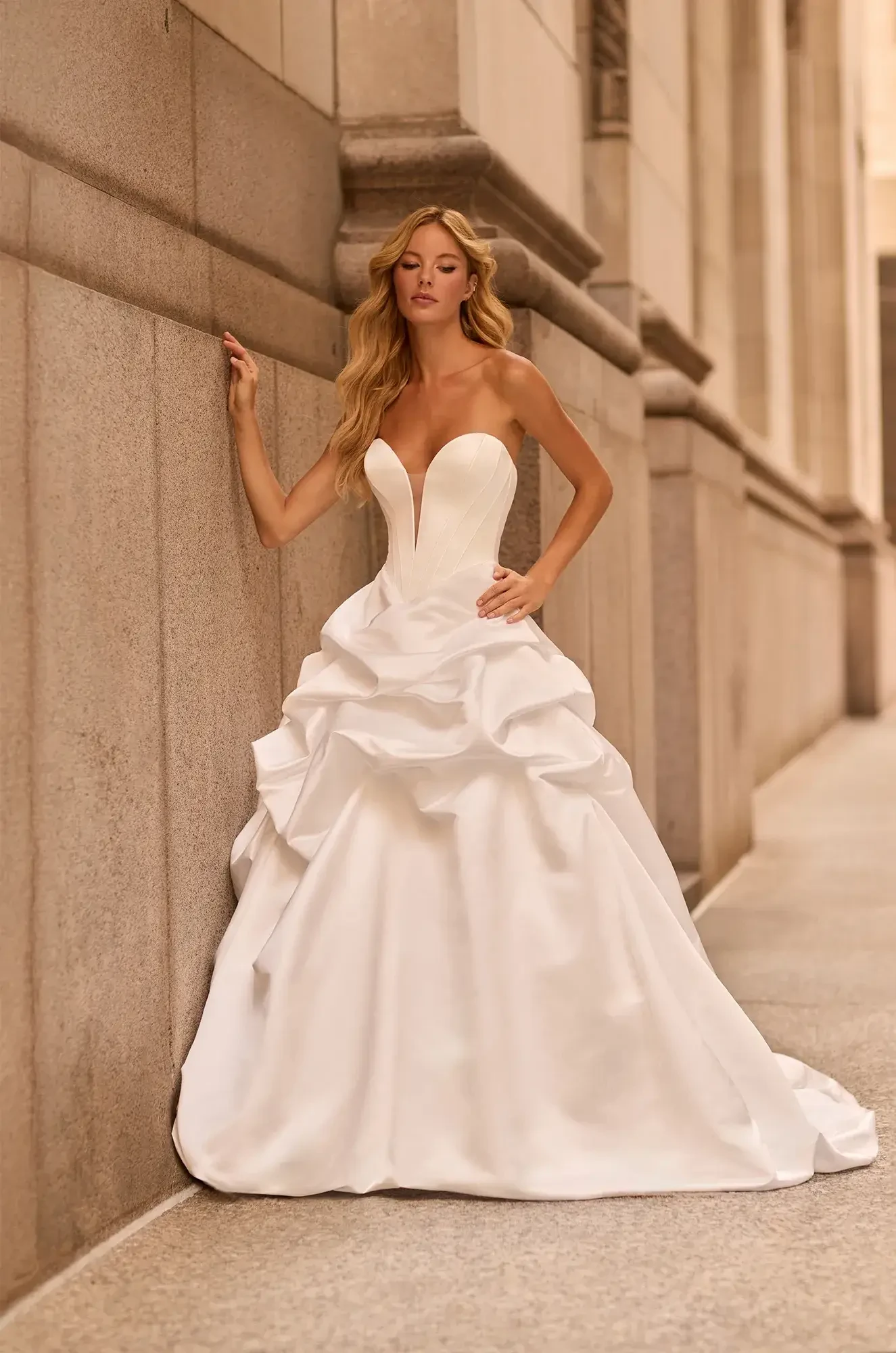 A woman in a white strapless wedding dress with a layered skirt, leaning against a wall.
