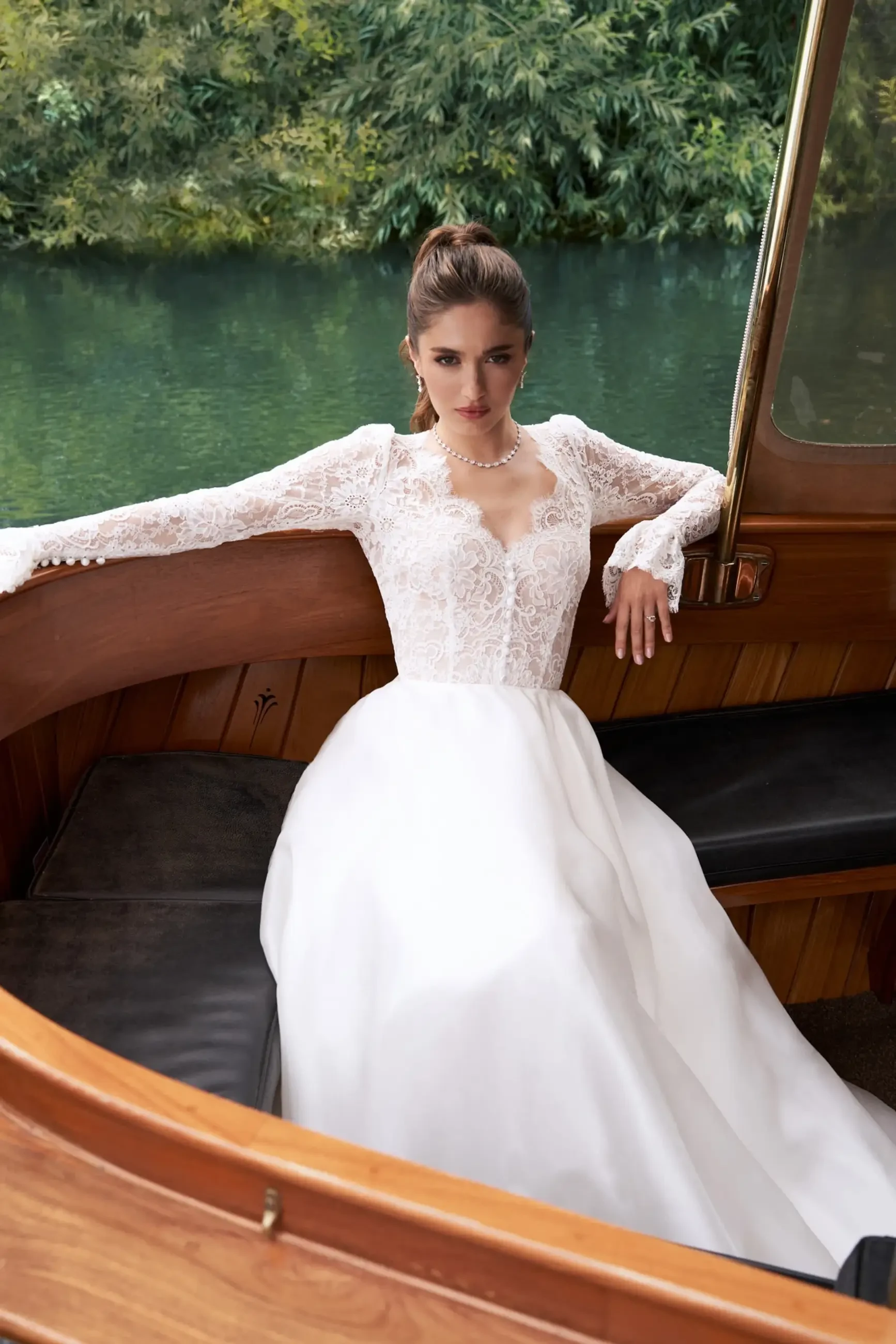 Bride in a lace long-sleeved gown sits in a wooden boat, surrounded by lush greenery and calm water, conveying serenity and elegance.