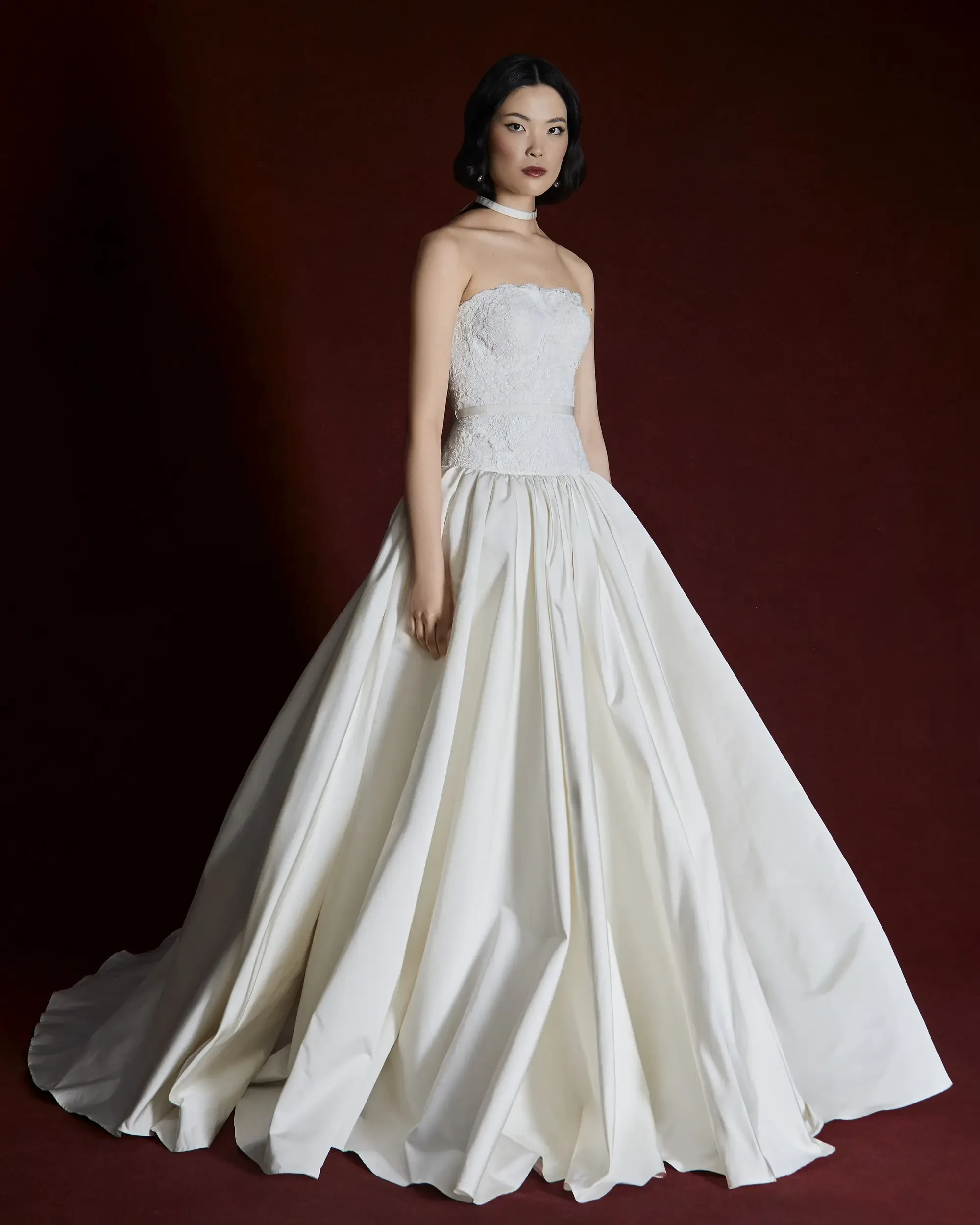 A model wearing a strapless white gown with a full skirt against a deep red background.