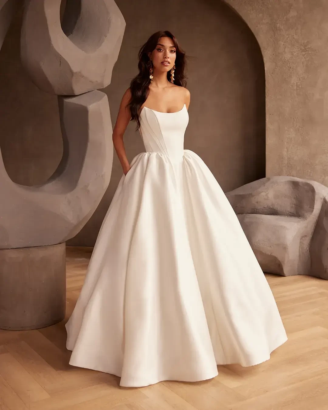 A woman in an elegant strapless white ball gown poses with hands in pockets. The room features abstract stone sculptures, creating a sophisticated atmosphere.