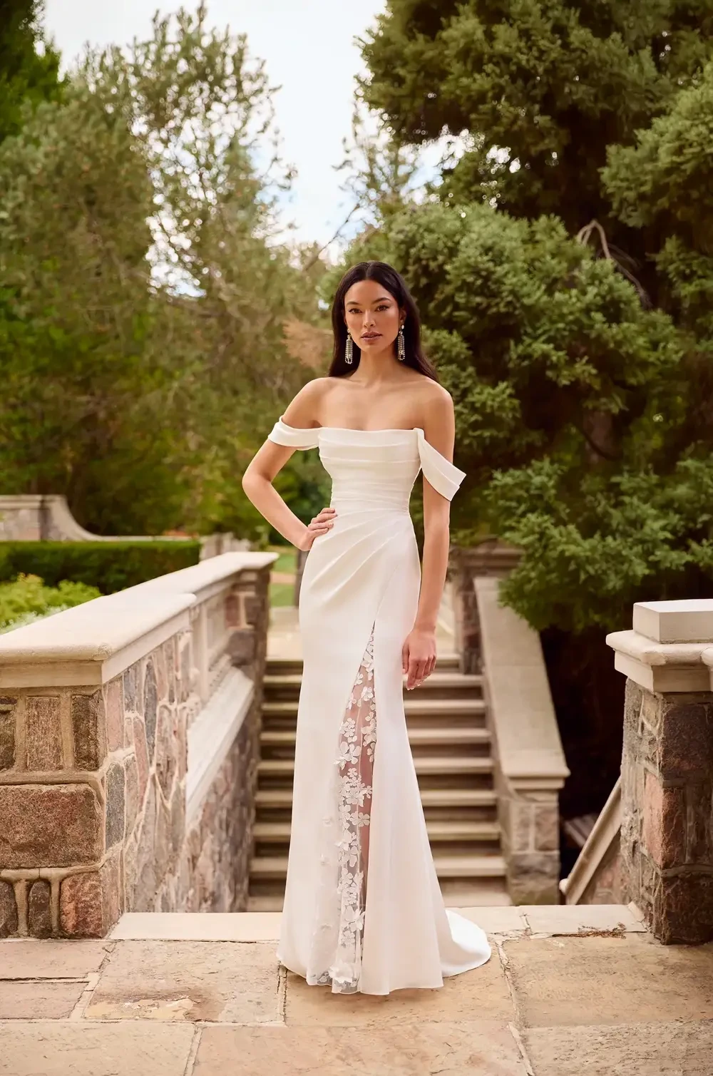 A woman in a white off-the-shoulder dress with a thigh-high slit stands on a stone pathway surrounded by greenery.