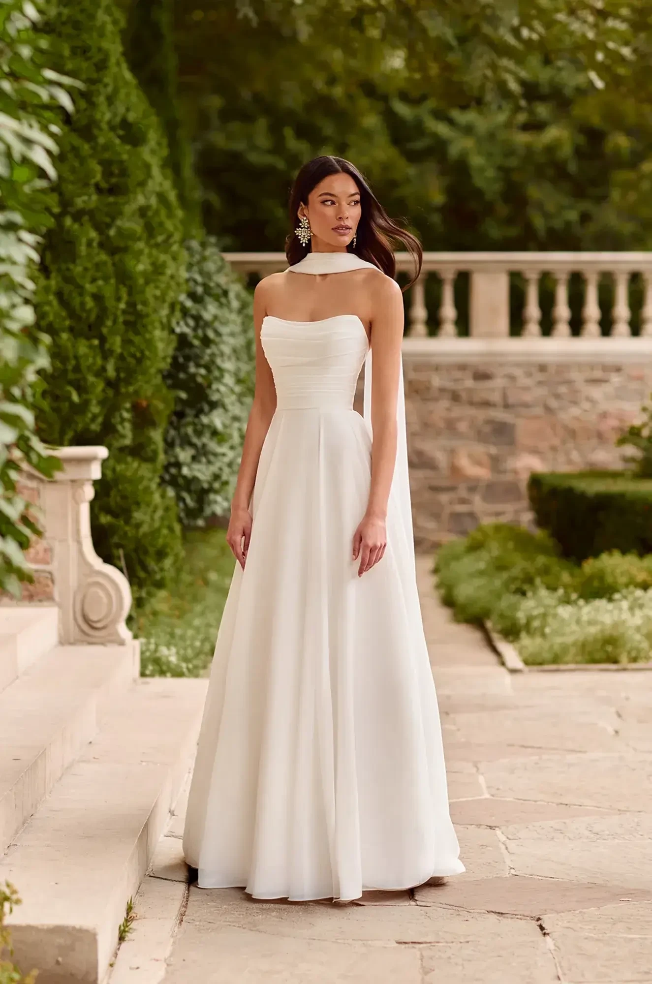A model wearing a white gown with a cape stands on steps in a garden setting.