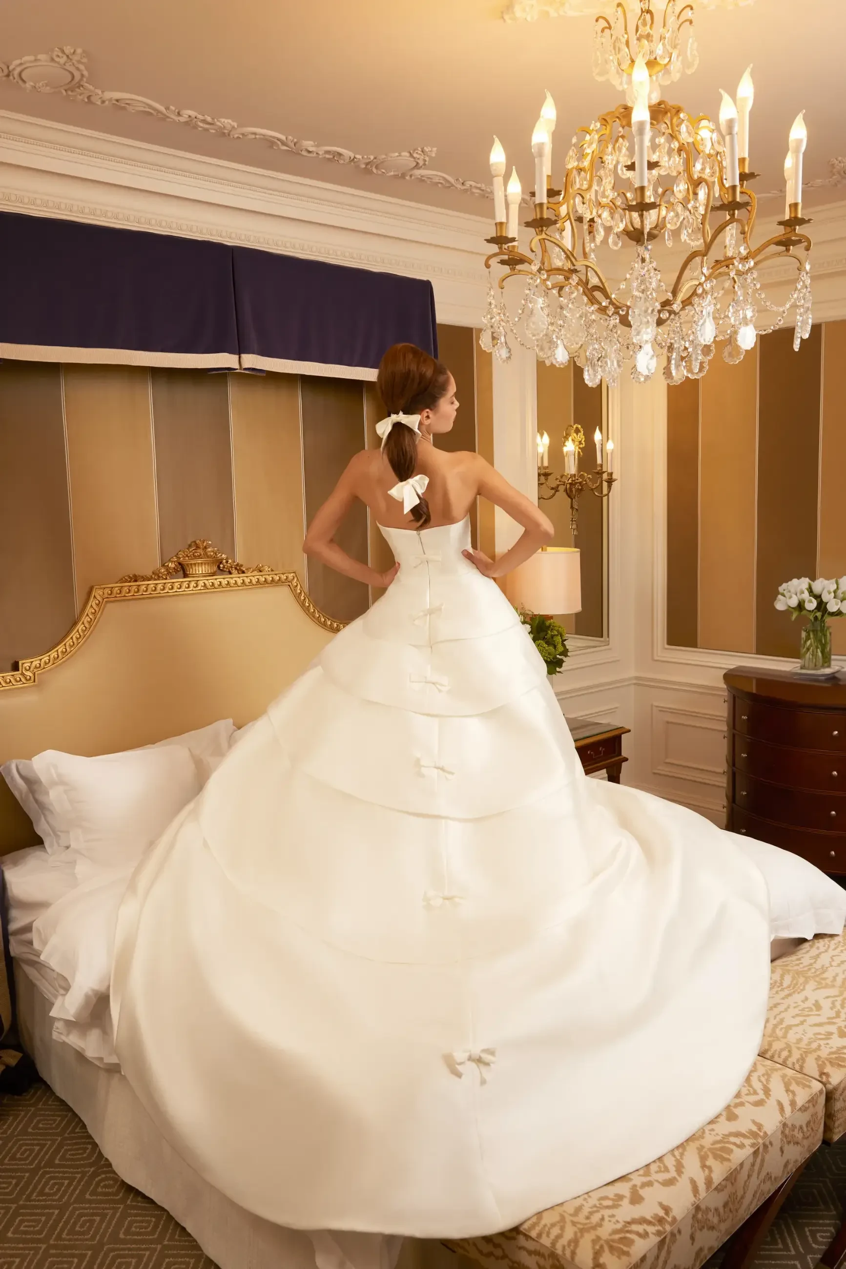 Bride in strapless gown with a voluminous skirt adorned with bows stands on a bed beneath an elegant chandelier, in a luxurious, ornate room.