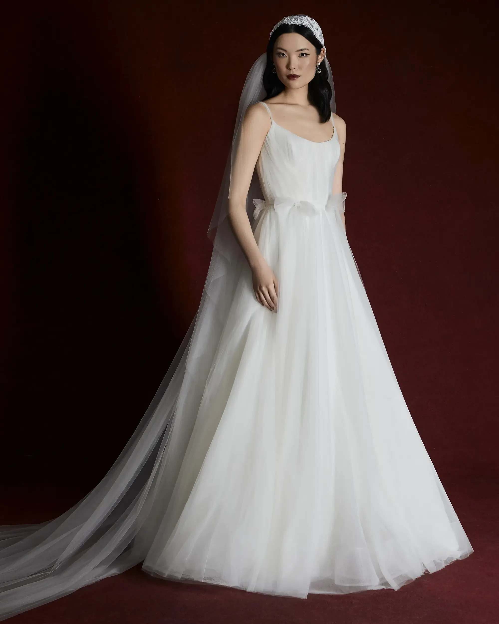 A model in a white wedding gown with a long veil, standing against a dark red background.